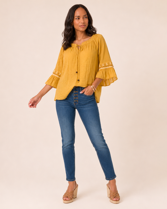 Amber Bloom Embroidery Peasant Top Close-up of a woman wearing a mustard yellow peasant top with a tassel tie neckline, ruffle sleeves, and decorative trim details on a beige background