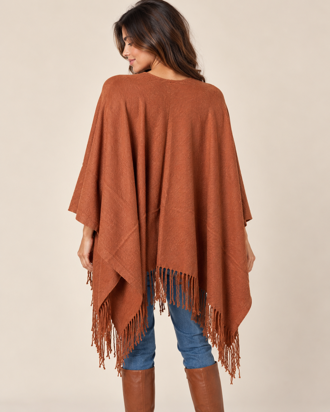 A woman shown from the back wearing a rust-brown fringed poncho over blue jeans and brown boots, against a plain beige studio background.