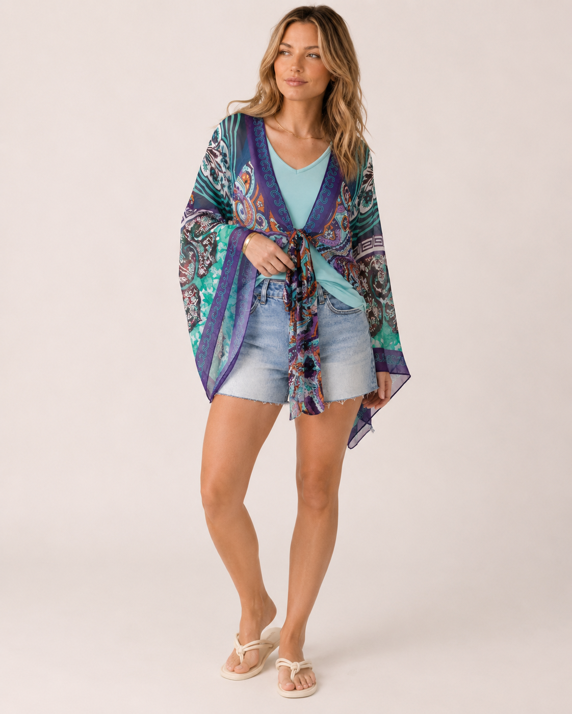Full-body view of a woman wearing a blue floral front-tie kimono over a blue V-neck tank top, light wash denim shorts, and brown sandals on a beige background