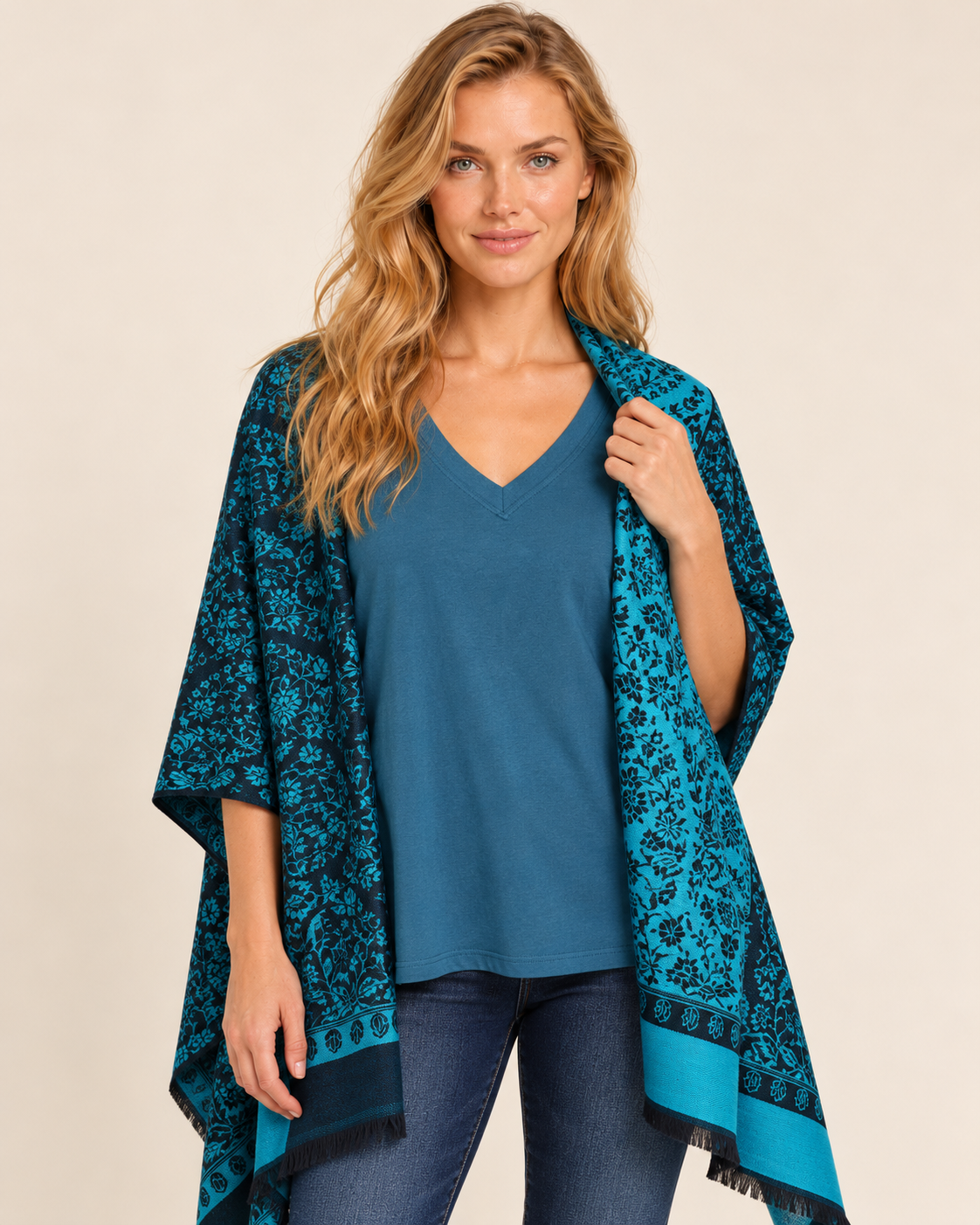 Cropped body front view of a woman wearing a teal and black Southwestern geometric-patterned poncho cardigan over a dark top, paired with dark blue skinny jeans and black ankle boots on a beige background.