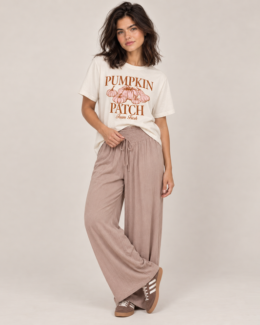 Pretty Little Pumpkin Patch Graphic Tee A woman wearing a cream-colored graphic t-shirt with a brown and pink 'Pumpkin Patch Farm Fresh' print, paired with light beige pants, against a neutral beige background.