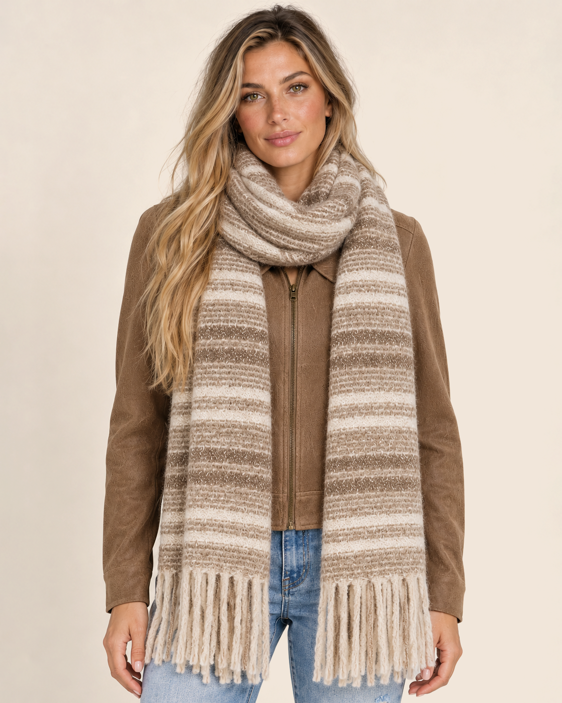 Full-body front view of a woman wearing a long brown, beige, and cream striped scarf with fringed ends over a brown top and light-wash blue jeans on a beige background.
