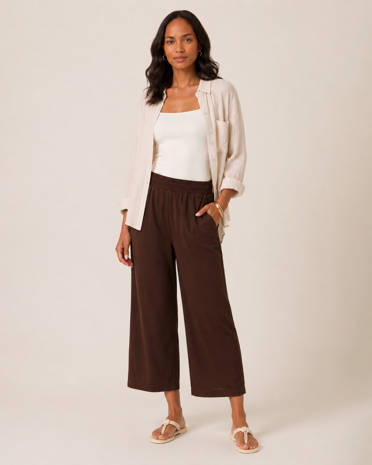 Front view of a woman wearing brown wide-leg pants, a white tank top, and a beige button-front shirt on a beige background