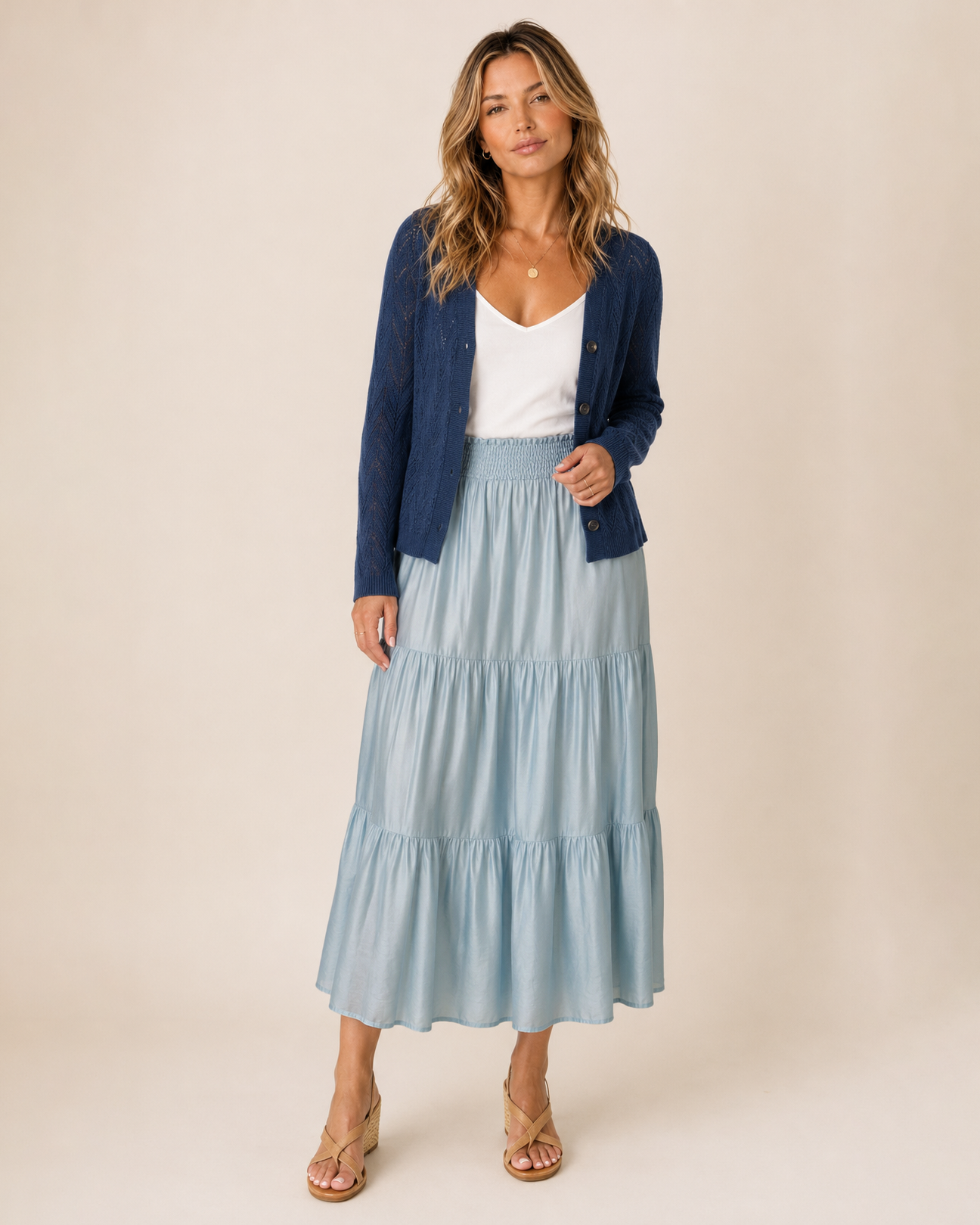A woman modeling a light blue tiered maxi skirt with a white top and a dark blue cardigan, standing against a solid beige studio background.