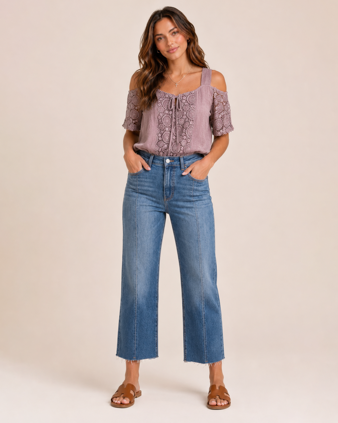 Full-body view of a woman wearing a mauve cold shoulder lace blouse, cropped blue jeans, and brown sandals on a beige background