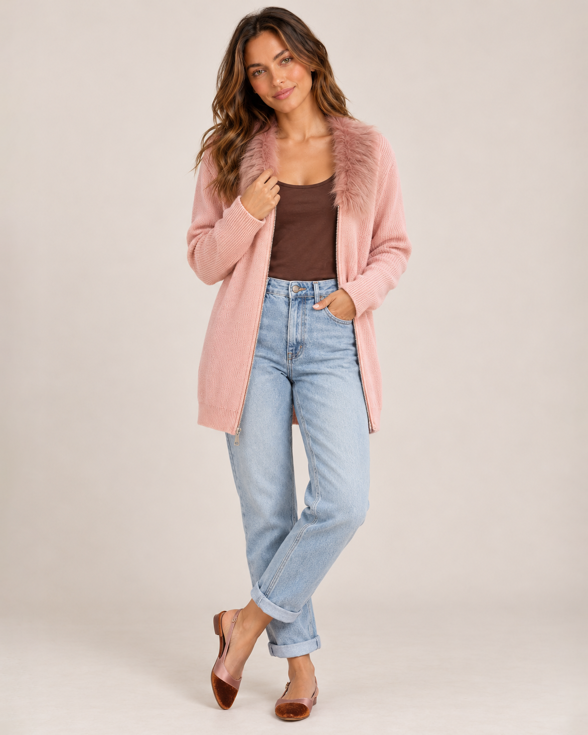 Full-body front view of a woman wearing a blush pink zip-front cardigan with a fluffy faux-fur collar over a brown fitted top, paired with light-wash blue jeans on a beige background.
