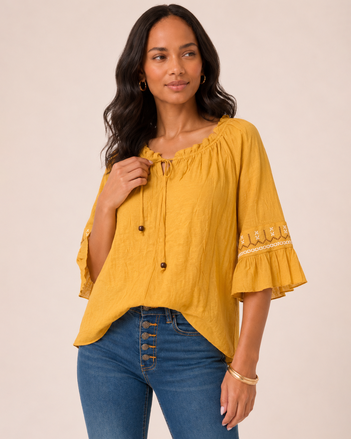 Close-up of a woman wearing a mustard yellow peasant top with a tassel tie neckline, ruffle sleeves, and decorative trim details on a beige background