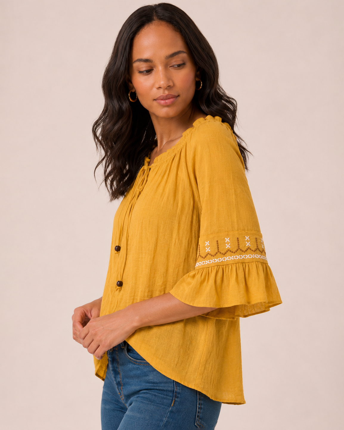 Side view of a woman wearing a mustard yellow peasant top with ruffle sleeves, decorative trim details, and blue jeans on a beige background