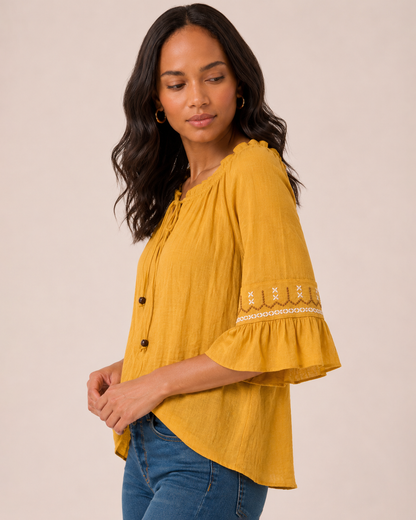 Side view of a woman wearing a mustard yellow peasant top with ruffle sleeves, decorative trim details, and blue jeans on a beige background