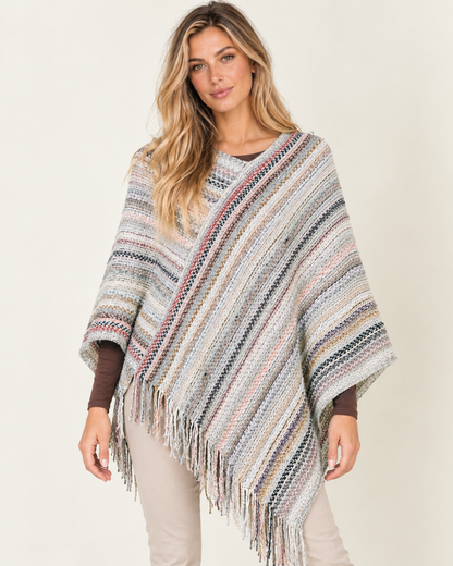 A woman wearing a multicolor striped knit poncho with fringe, styled over a brown long-sleeve top and beige pants, against a plain light beige background.