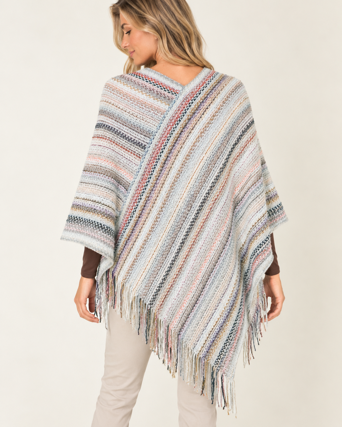 A woman wearing a multicolor striped knit poncho with fringe, shown from the back against a plain light beige studio background.