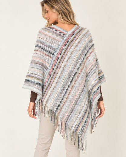 A woman wearing a multicolor striped knit poncho with fringe, shown from the back against a plain light beige studio background.