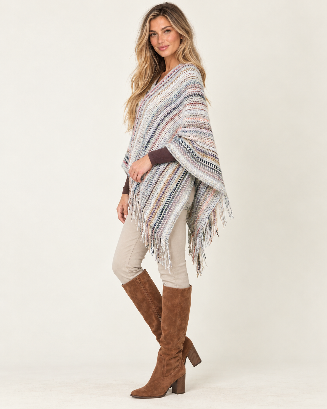 A woman modeling a multicolor striped knit poncho with fringe, worn over a dark brown long-sleeve top and paired with beige pants and brown knee-high boots, against a plain light background.