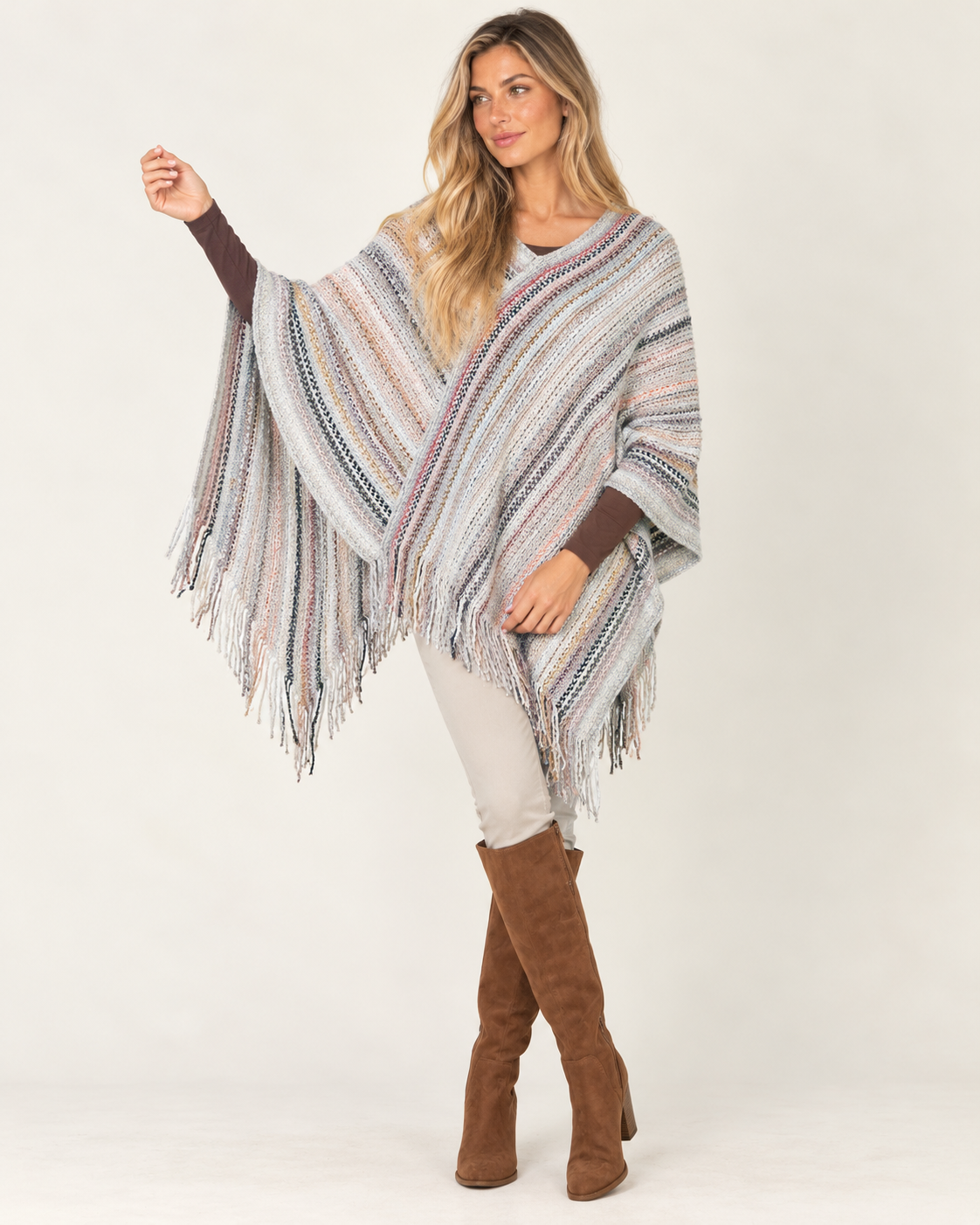 A woman modeling a striped knit poncho with fringe, worn over a white top and paired with brown suede knee-high boots, against a plain light background.