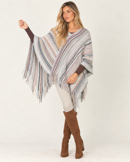 A woman modeling a striped knit poncho with fringe, worn over a white top and paired with brown suede knee-high boots, against a plain light background.