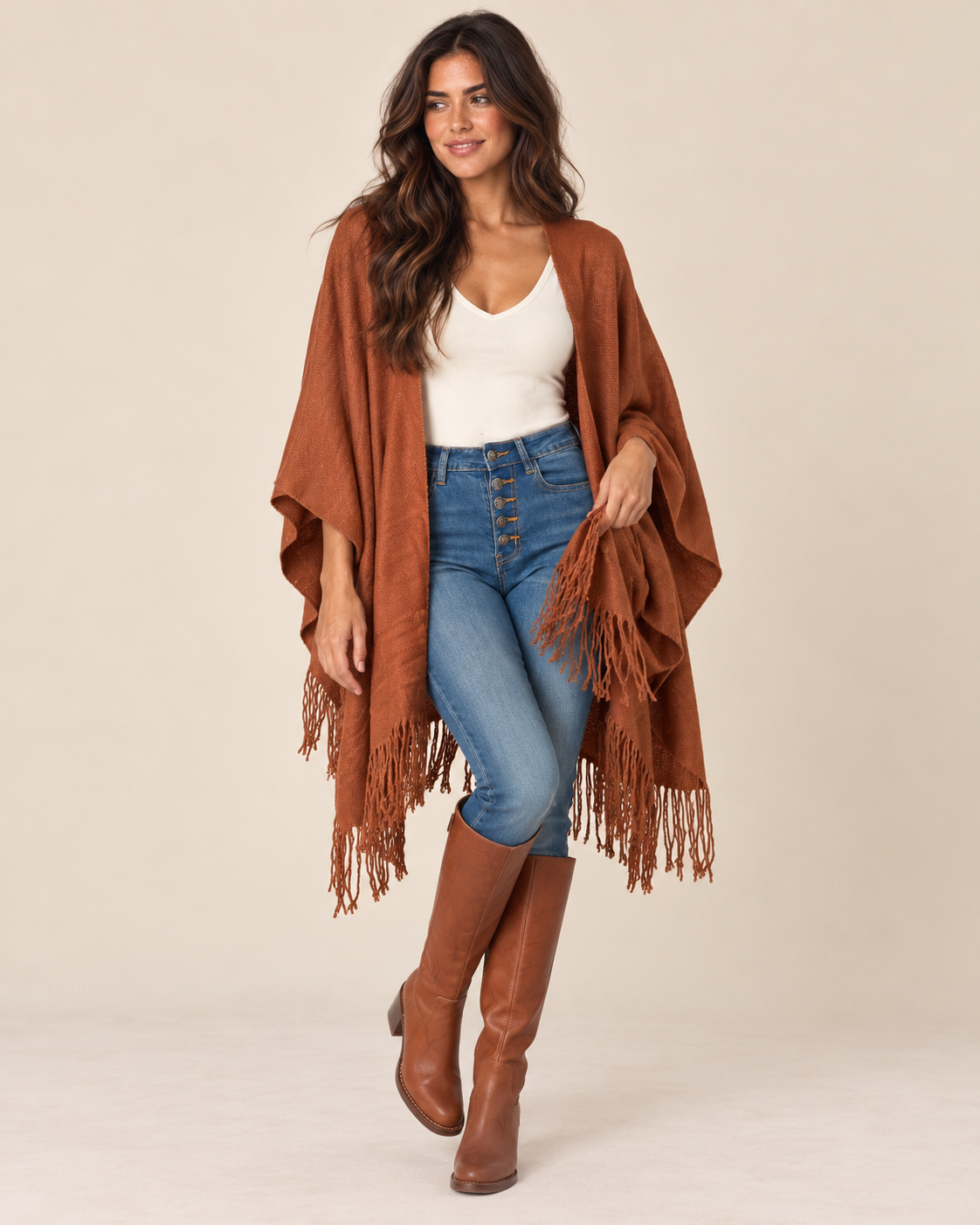 A woman modeling a rust-brown fringed poncho over a white tank top, blue jeans, and brown knee-high boots, set against a plain beige studio background.