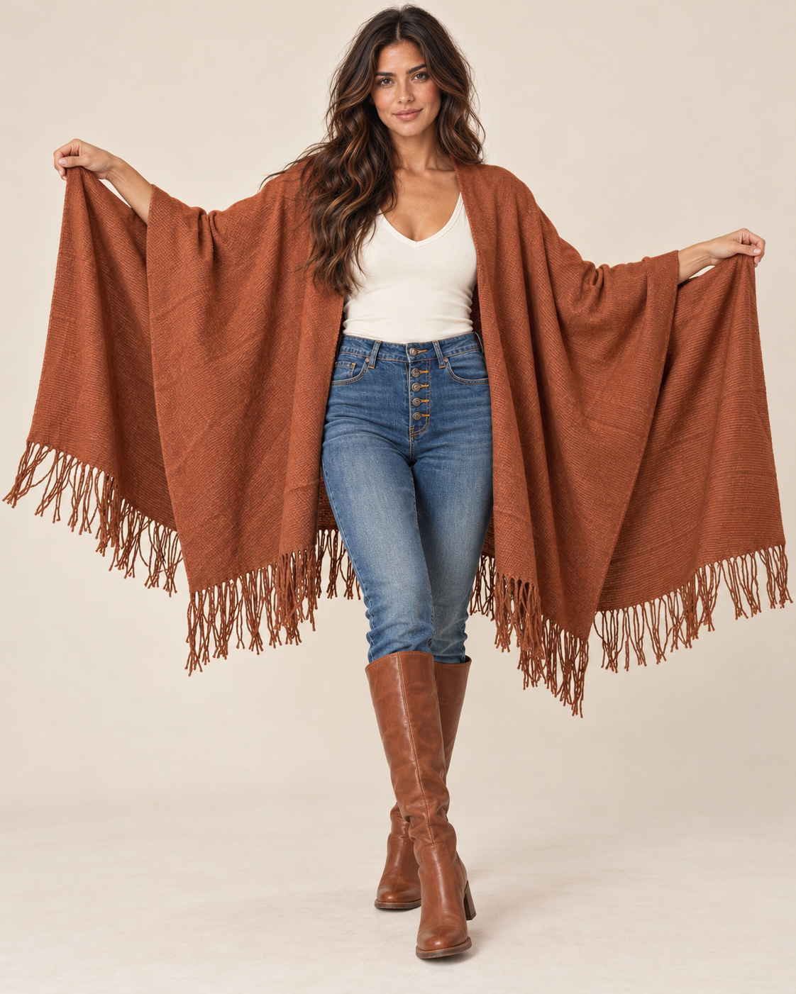 A woman modeling a rust-brown fringed poncho with wide sleeves, worn over a white tank top and blue jeans, against a plain beige studio background.