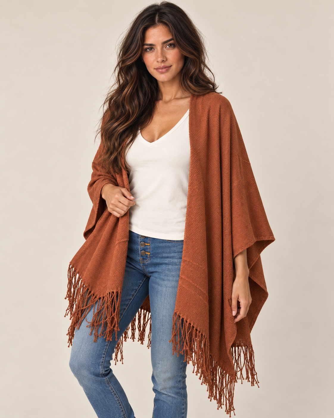 A woman modeling a rust-brown fringed poncho or shawl over a white V-neck top and blue jeans, photographed against a plain light beige studio background.