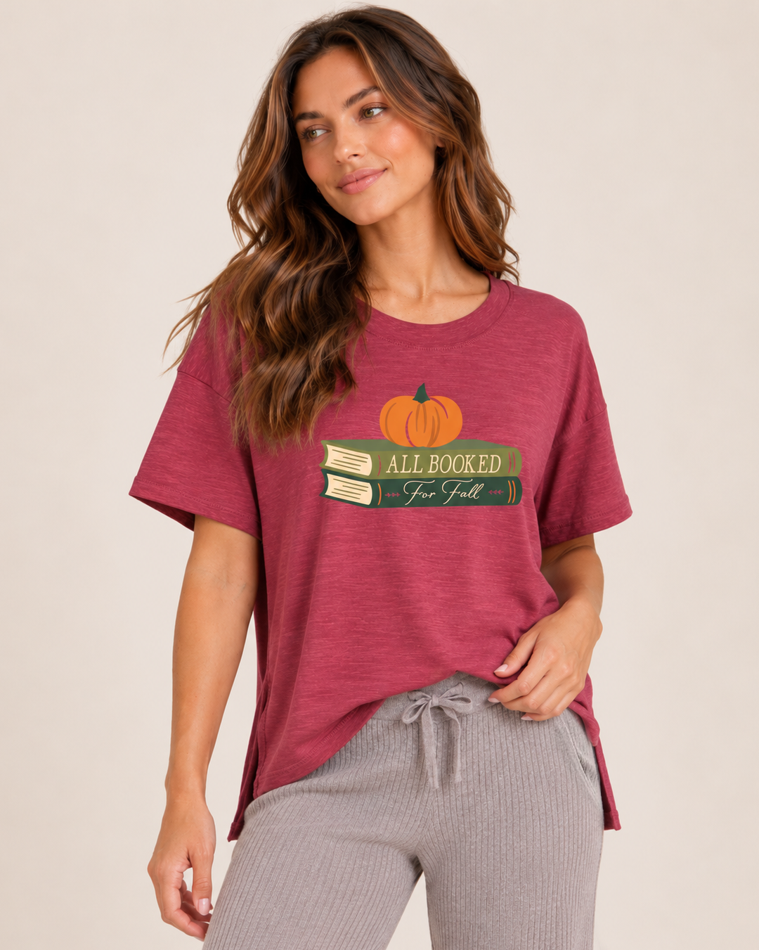 A woman wearing a heathered dusty rose short-sleeve t-shirt with a graphic of a pumpkin on stacked books that read 'All Booked for Fall", paired with gray drawstring pants, against a solid beige background.