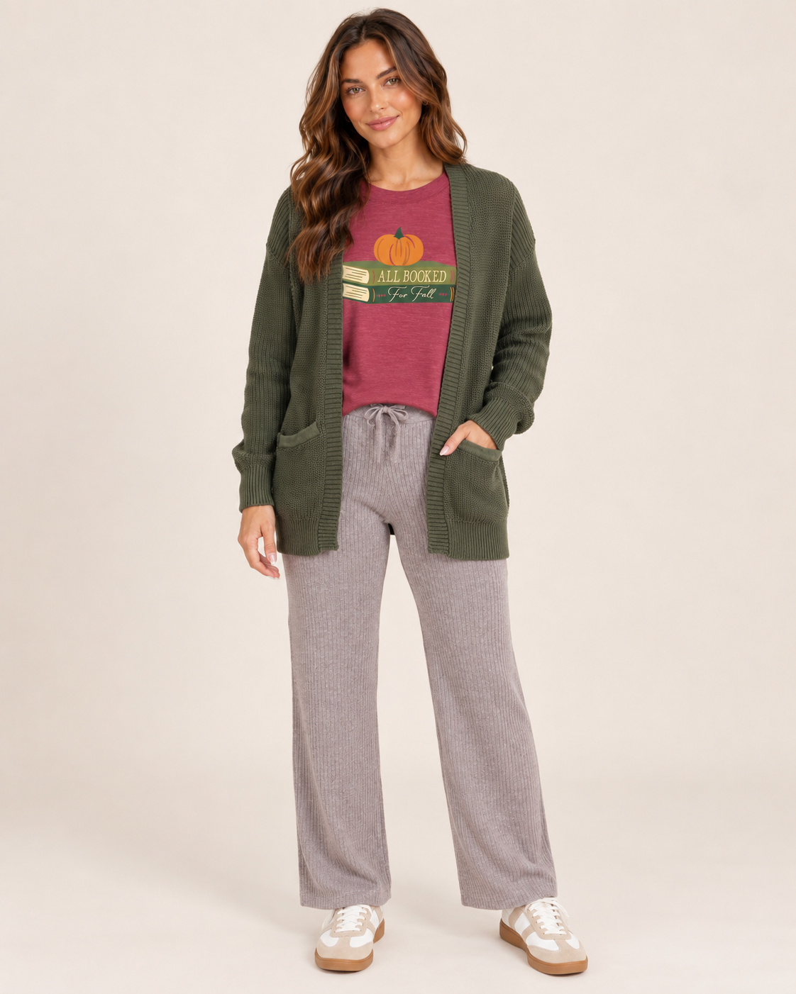 A woman modeling a cozy fall outfit consisting of an olive green open-front cardigan, a burgundy graphic t-shirt, heather gray drawstring lounge pants, and white sneakers, set against a plain beige studio background.