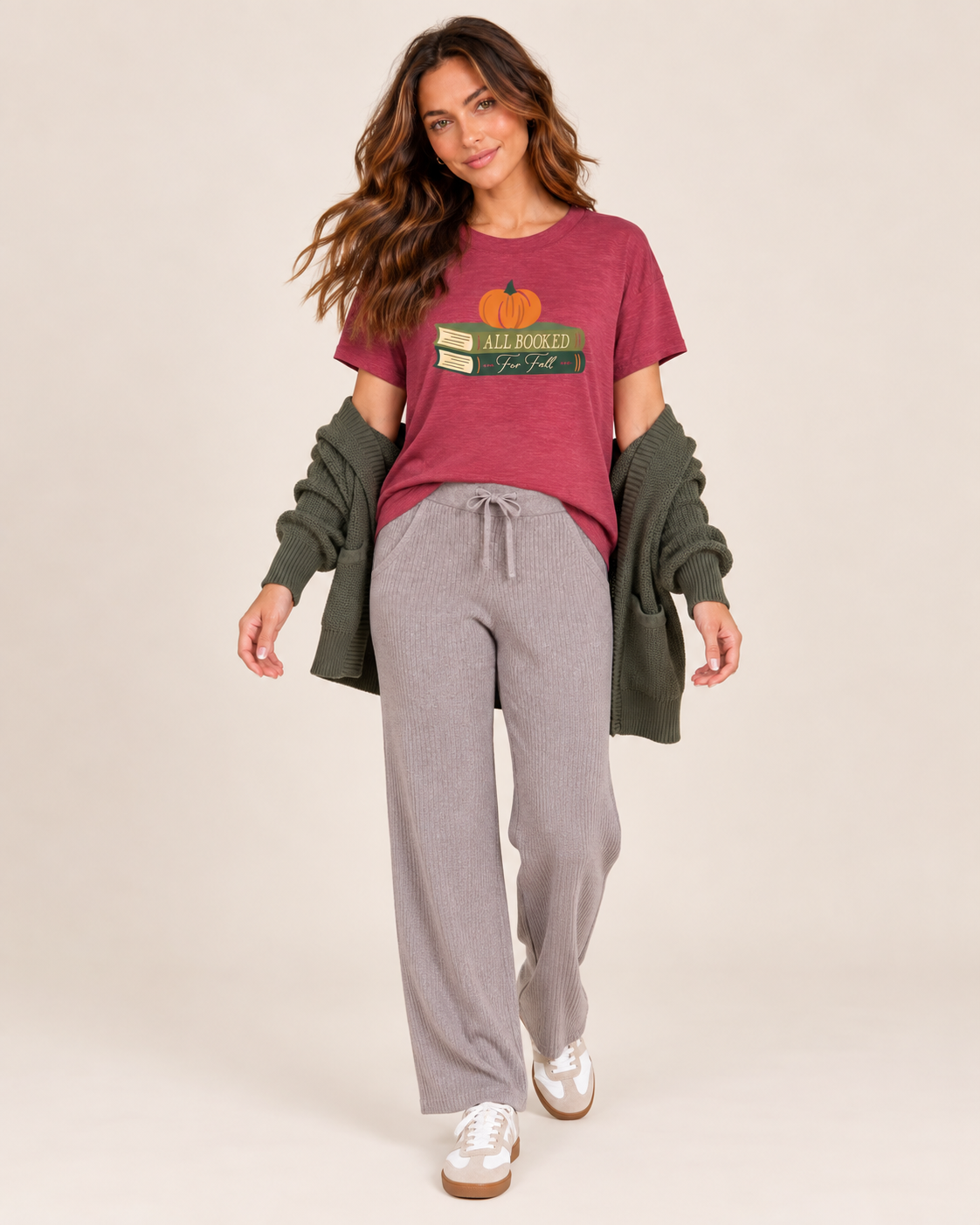 A woman modeling a cozy fall-themed outfit consisting of a burgundy graphic t-shirt with a pumpkin and books design, gray drawstring sweatpants, an olive green cardigan, and white sneakers, set against a plain beige studio background.