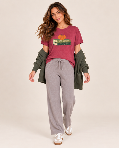 A woman modeling a cozy fall-themed outfit consisting of a burgundy graphic t-shirt with a pumpkin and books design, gray drawstring sweatpants, an olive green cardigan, and white sneakers, set against a plain beige studio background.