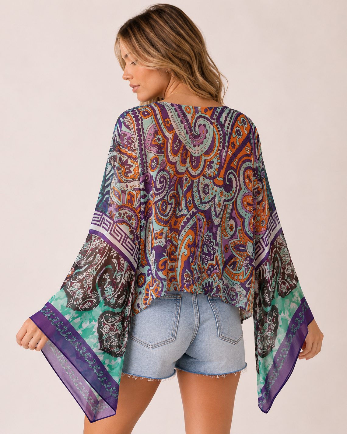 Back view of a woman wearing a blue floral front-tie kimono over a blue V-neck tank top and light wash denim shorts on a beige background