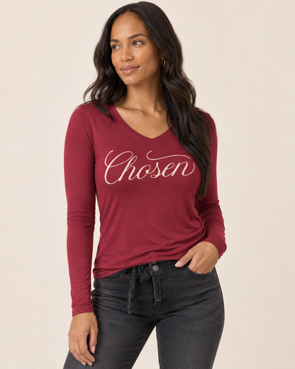 A woman modeling a long-sleeve burgundy V-neck t-shirt with 'Chosen' printed in white script across the chest, paired with dark gray jeans against a solid beige background.