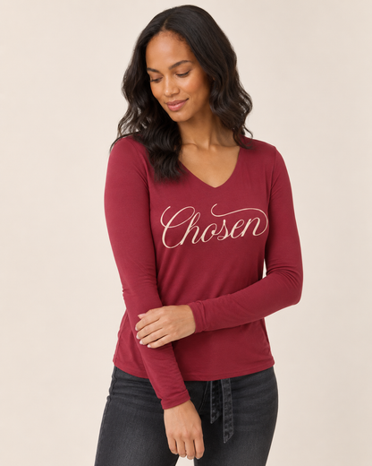 A woman wearing a burgundy long-sleeve V-neck top with 'Chosen' printed in white script across the chest, paired with dark gray jeans, against a solid beige background.