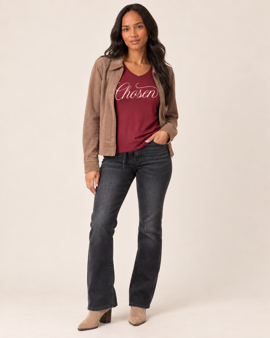 A woman modeling a brown suede jacket over a burgundy t-shirt with 'Chosen' printed in white script, paired with dark gray flare jeans and beige ankle boots, against a solid light beige background.