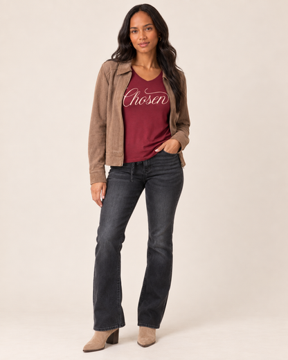 A woman modeling a brown suede jacket over a burgundy t-shirt with 'Chosen' printed in white script, paired with dark gray flare jeans and beige ankle boots, against a solid light beige background.