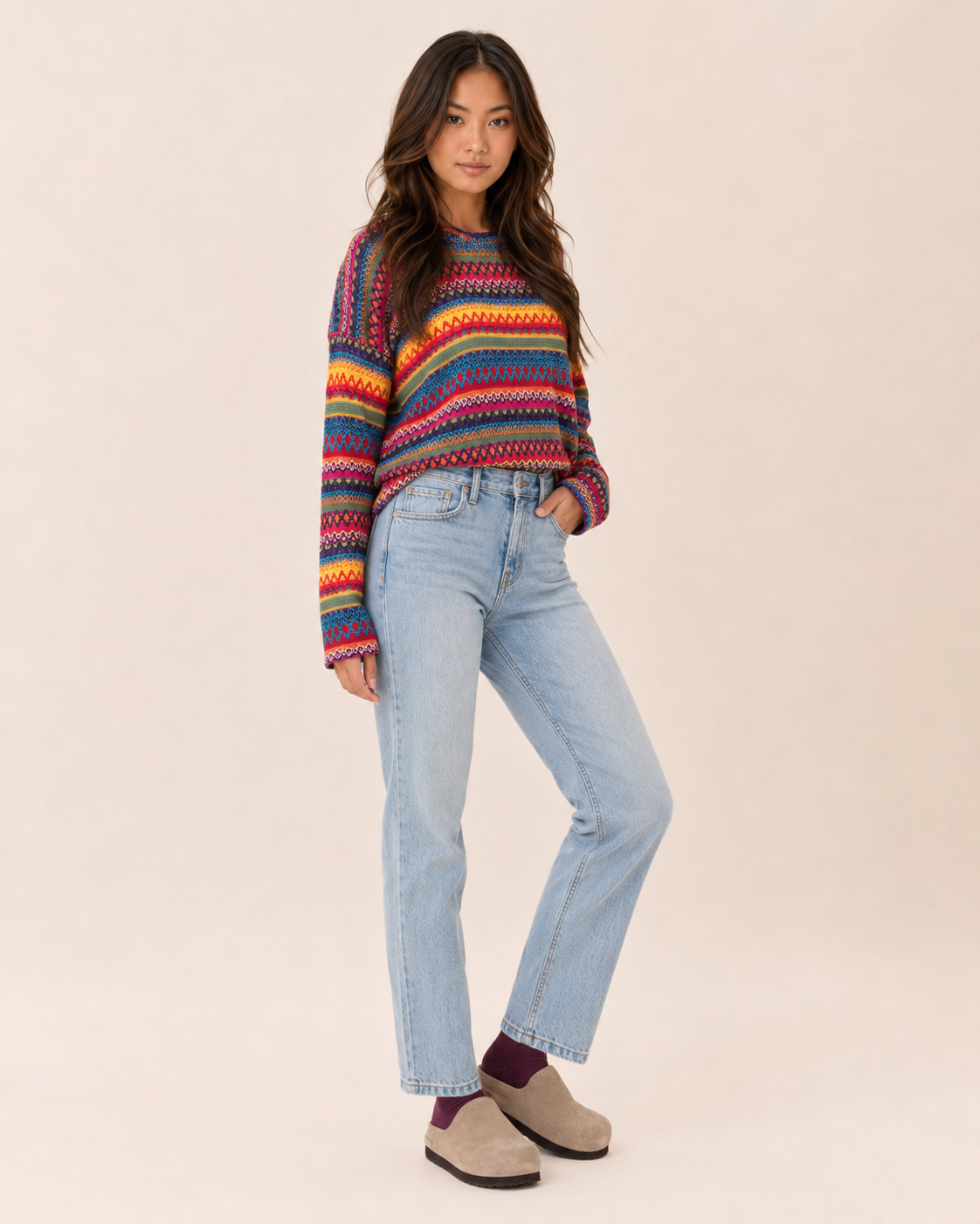 Full-body view of a woman wearing a multicolor striped knit pullover sweater with light wash straight-leg jeans and tan slip-on shoes on a beige background