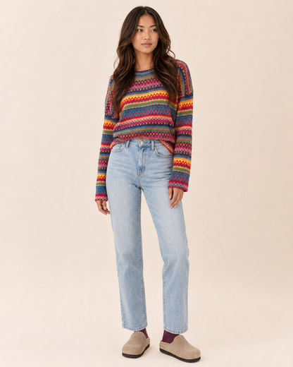 Full-body view of a woman wearing a multicolor striped knit pullover sweater with light wash straight-leg jeans, burgundy socks, and tan slip-on shoes on a beige background