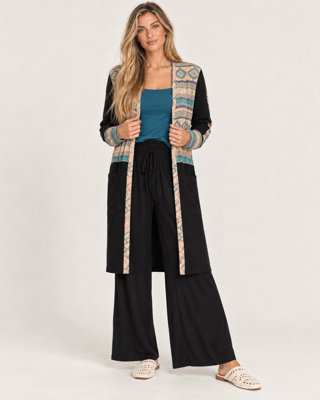 Full-body front view of a woman wearing a long black maxi cardigan with multicolor geometric patterned trim, a white top, and light-wash flared jeans on a beige background.