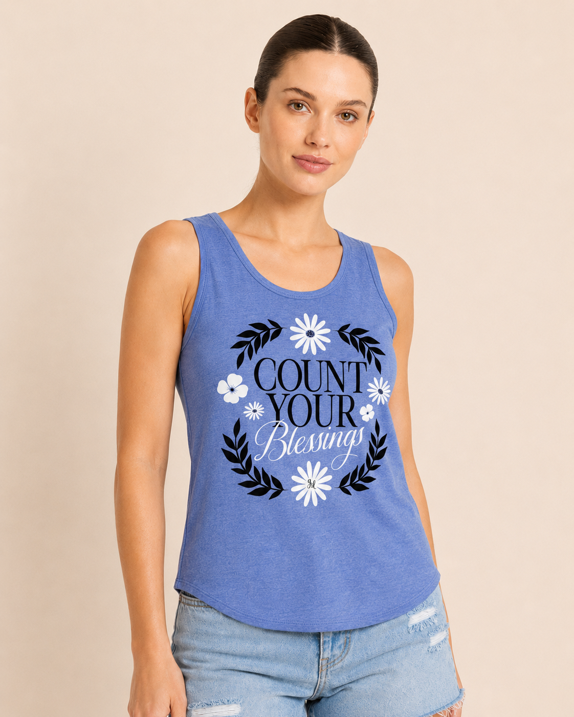 Close-up of a woman wearing a blue graphic tank top featuring a "Count Your Blessings" floral design and light wash denim shorts on a beige background