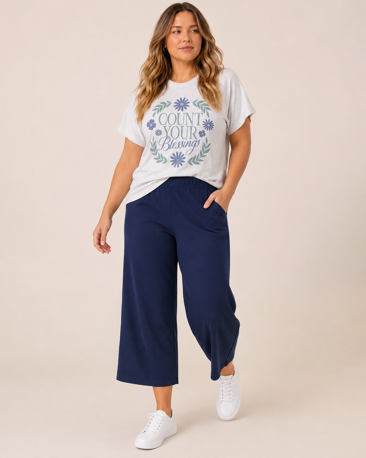 Full-body view of a woman wearing a white graphic tee featuring a "Count Your Blessings" floral design, navy wide-leg pants, and white sneakers on a beige background