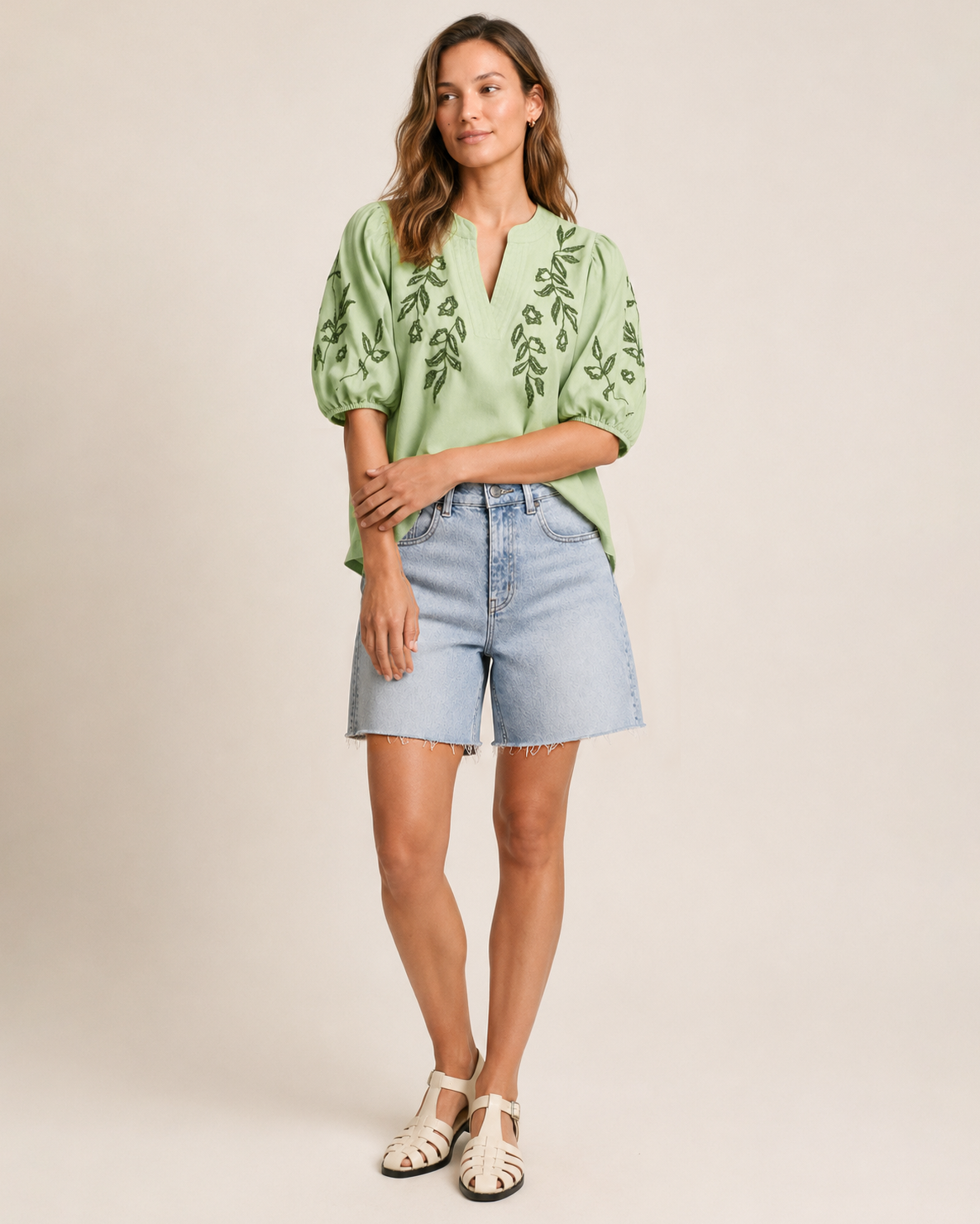 Full-body view of a woman wearing a sage embroidered blouse with puff sleeves and light wash denim shorts on a beige background