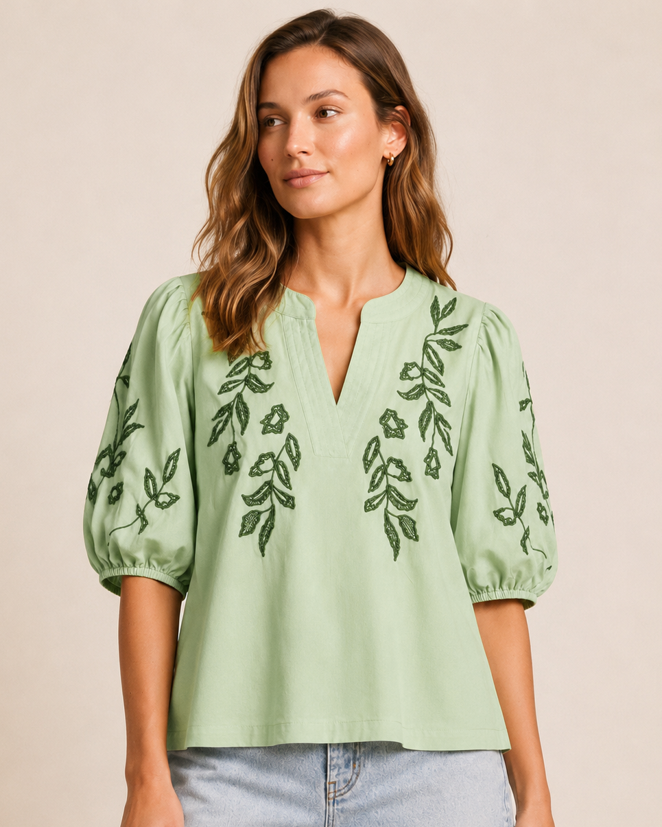 Front view of a woman wearing a sage embroidered blouse with puff sleeves and light wash denim shorts on a beige background