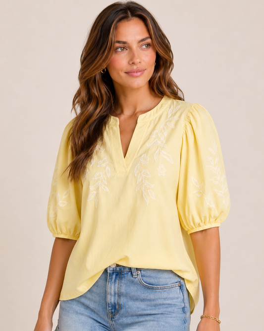 Close-up of a woman wearing a sunshine yellow embroidered blouse with puff sleeves and light wash denim shorts on a beige background