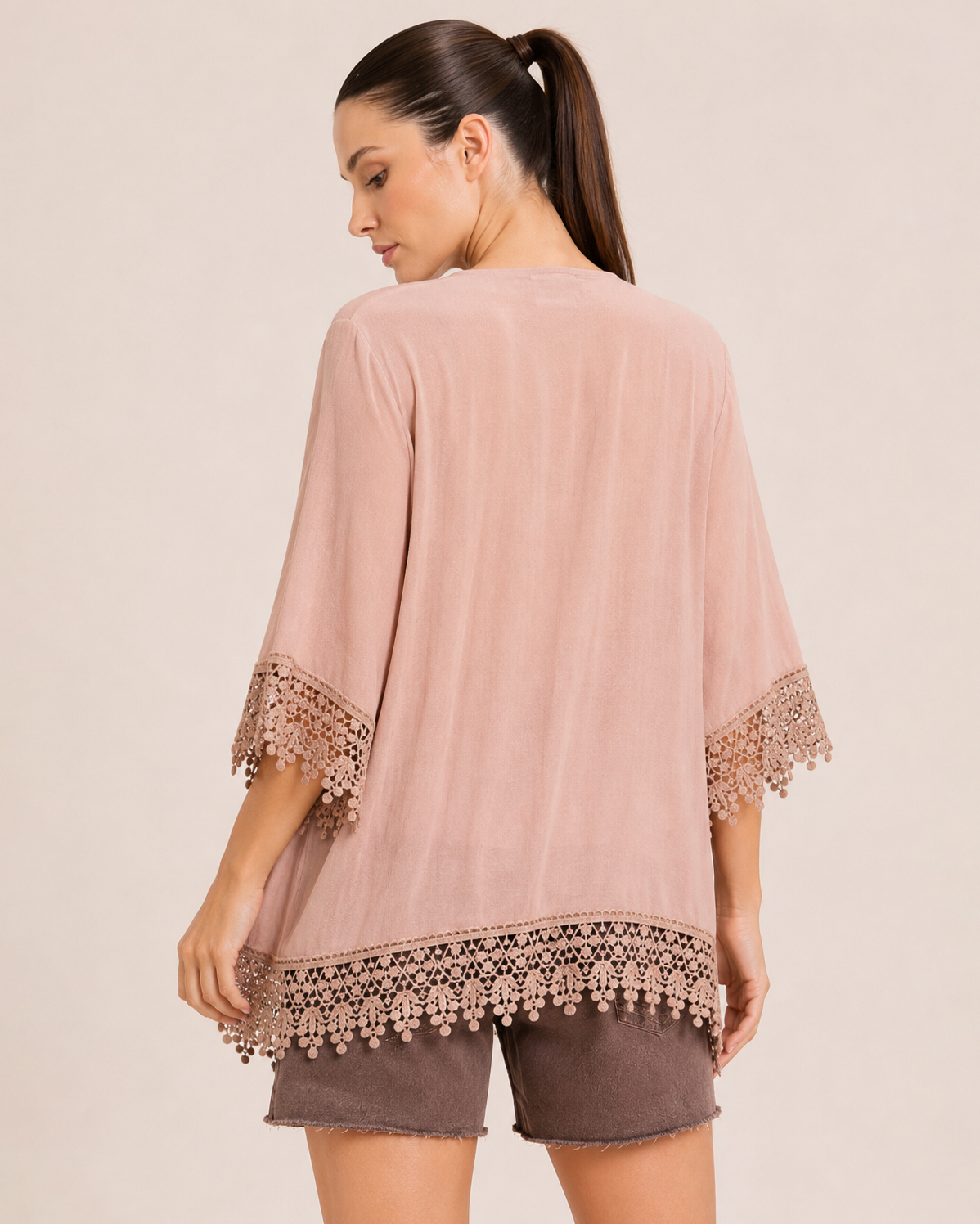 Back view of a woman wearing a blush lace trim kimono over a white V-neck tank top and brown distressed shorts on a beige background
