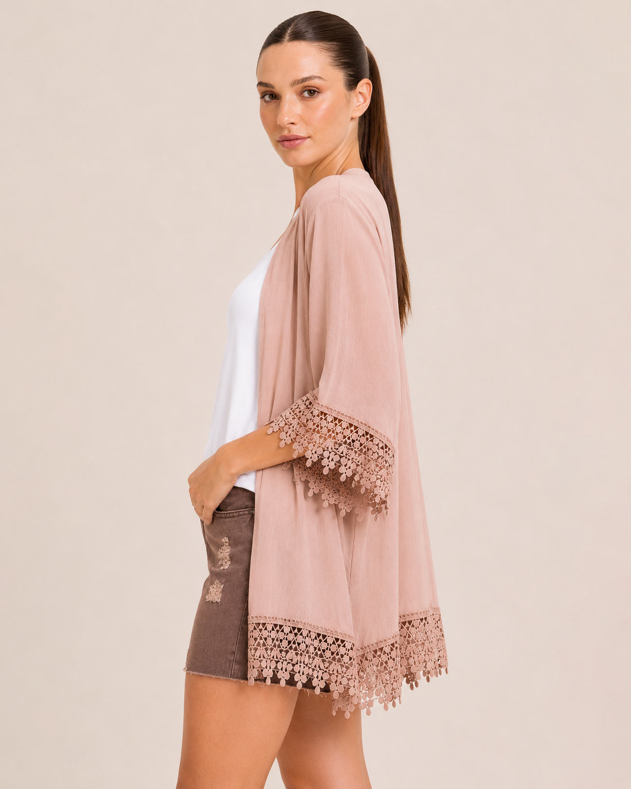 Side view of a woman wearing a blush lace trim kimono over a white V-neck tank top and brown distressed shorts on a beige background