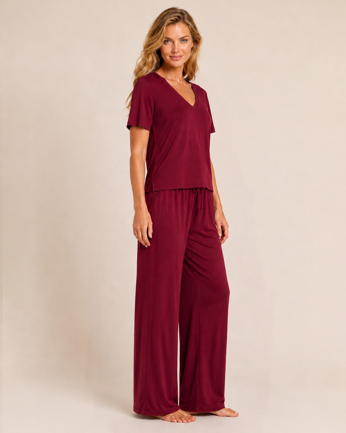 Full-body view of a woman wearing cabernet pajama pants with a matching V-neck top on a beige background