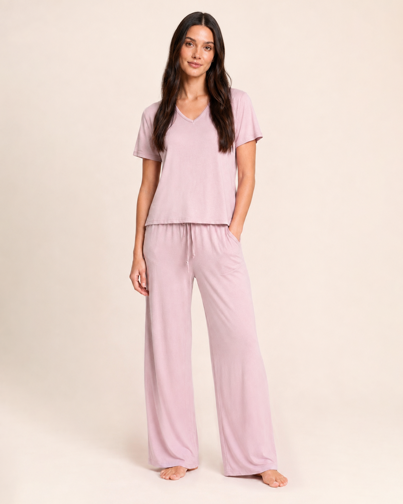 A woman modeling a two-piece pajama set in a dusty pink color, consisting of a short-sleeve V-neck top and matching wide-leg pants, against a solid light beige background.