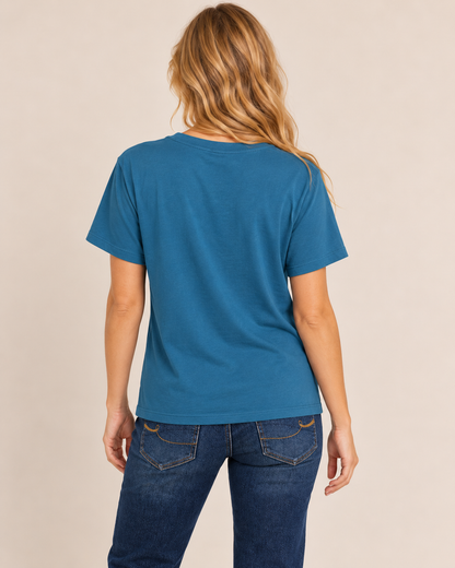 Back view of a woman wearing a teal blue short-sleeve top with dark blue jeans on a beige background.