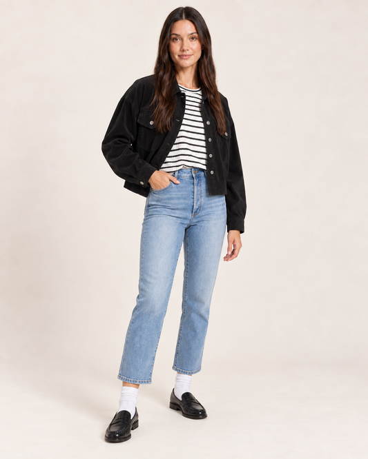 Cropped front view of a woman wearing an open black button-front cardigan over a black and white striped top, paired with light wash jeans on a beige background.