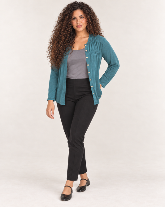 Full-body front view of a woman wearing black ankle-length leggings with a teal cardigan and black Mary Jane shoes on a beige background