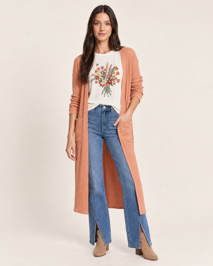 A woman modeling a long, ribbed peach cardigan over a cream graphic tee and blue jeans, standing against a plain light beige studio background.