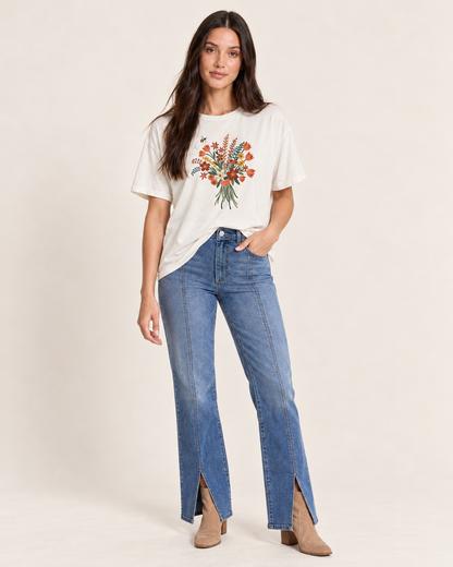 A woman modeling a white graphic t-shirt with a colorful floral print, paired with high-waisted blue jeans featuring front slits and light brown ankle boots, against a solid beige studio background.
