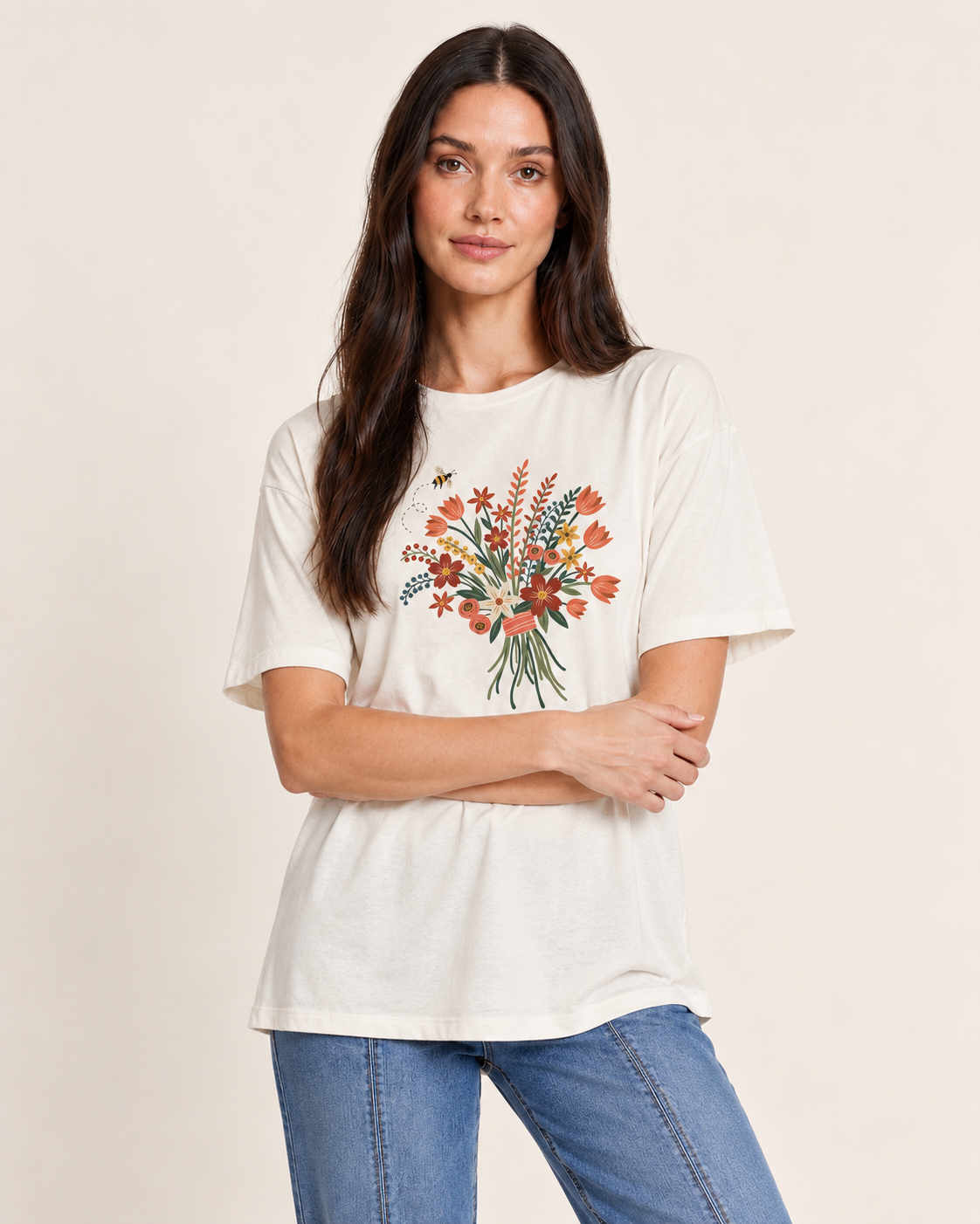 A woman modeling a cream-colored short-sleeve t-shirt with a colorful floral bouquet graphic on the front, paired with blue jeans, against a solid light beige background.