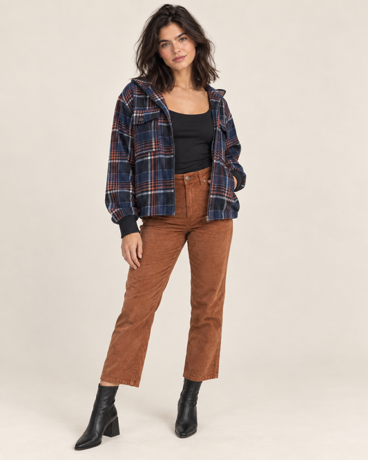 Cropped front view of a woman wearing a black spaghetti-strap camisole with brown straight-leg pants and a multicolor plaid overshirt on a beige background